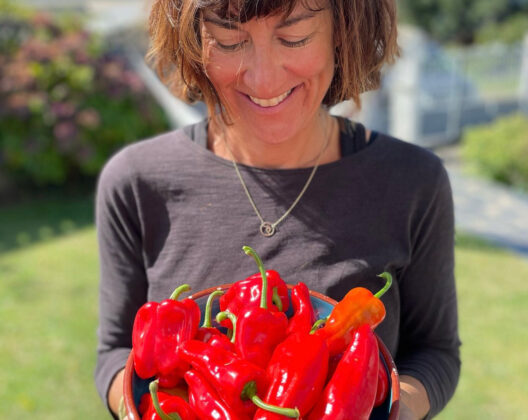 Smiling woman holding a bowl of fresh red bell peppers outdoors on a sunny day.